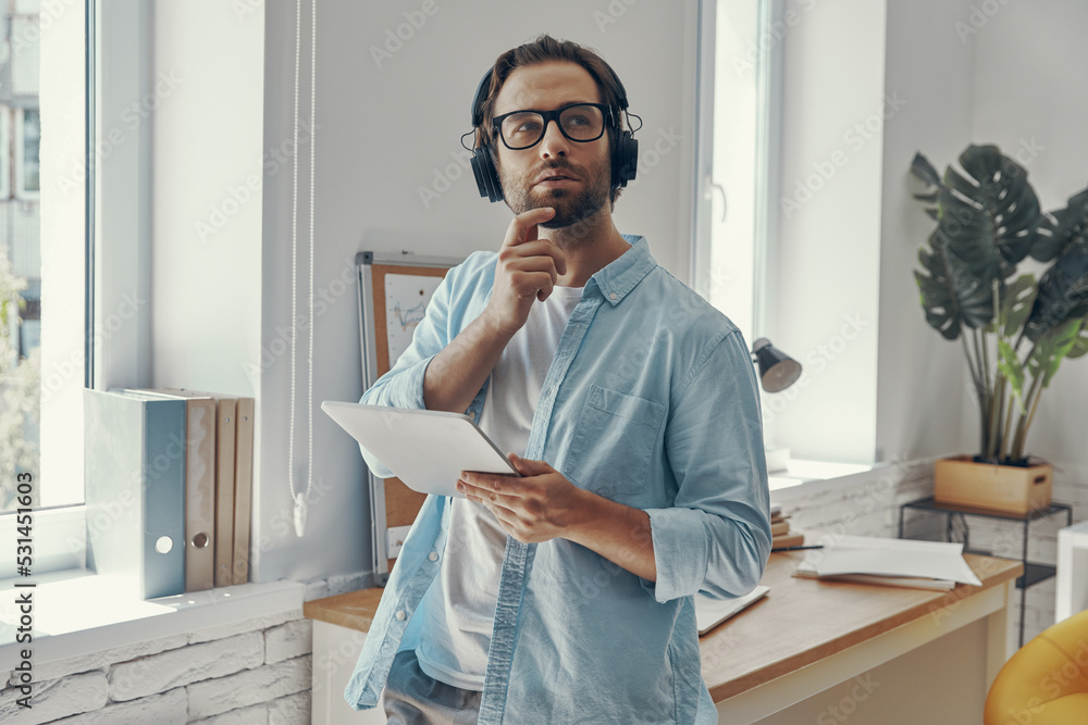 Fototapeta premium Thoughtful young man in headphones holding digital tablet while standing in the office