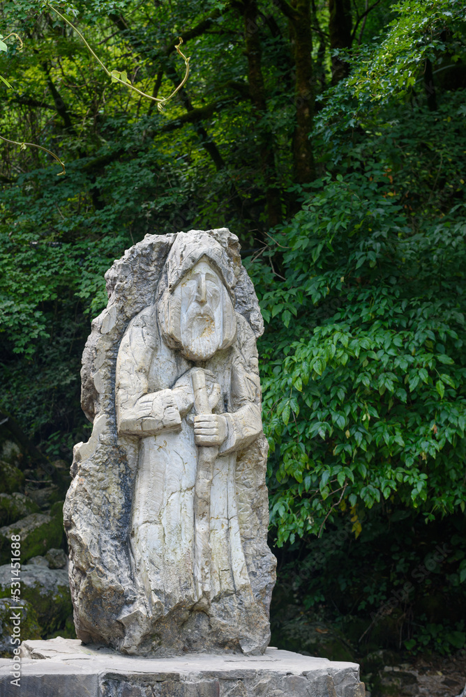 Stone statue standing among green trees in park symbolises old forest dwellers. Vintage sculpture attracts tourists attention among plants