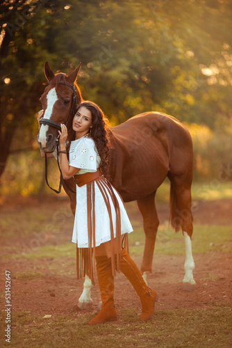 portrait of a young beautiful woman with curly hair who is holding a brown horse. Walking with the animals on the farm. Horse riding. boho style rider © Daria