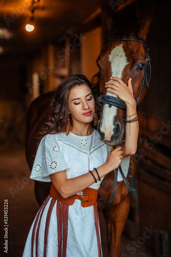 young woman in a white dress and a fringed belt holds a horse in a stall at the stable