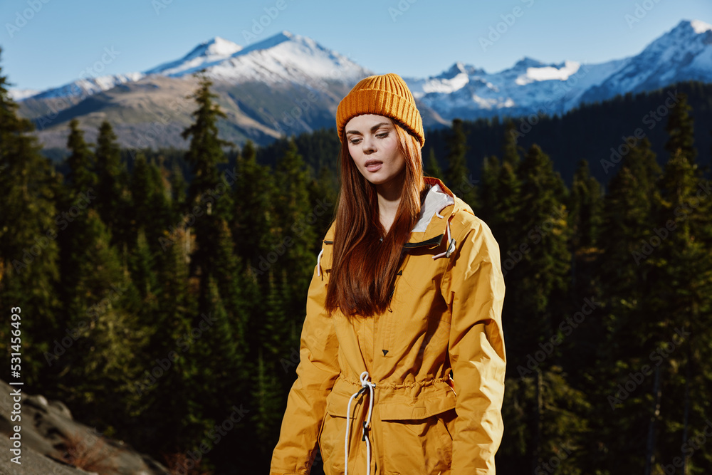 Woman in a yellow raincoat with red hair in a hike stands against the backdrop of the mountains in a yellow cap in the fall sunset light cute smile happiness