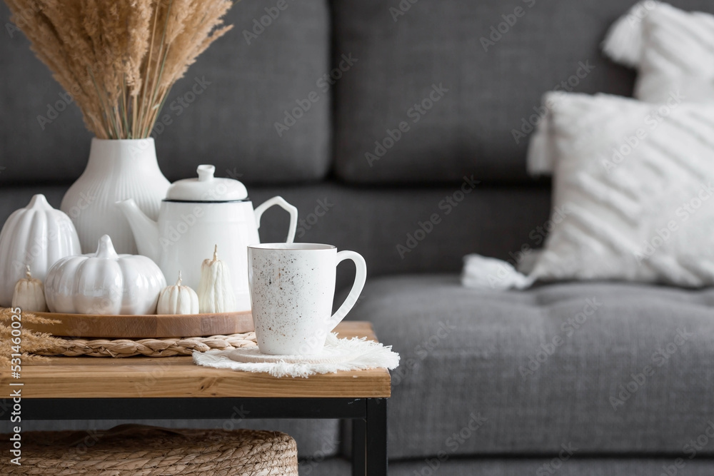 Still-life. Dried pampas grass in a vase, white ceramic pumpkins, a teapot, a cup and pumpkin-shaped candles on a coffee table in the home interior of the living room. Cozy autumn concept.