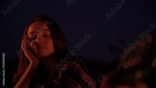 Young urban woman sit near the fire in the evening. Beautiful carefree trendy girl at sunset near the bonfire on the empty black sand tropical beach. Hipster young woman in trendy stylish clothes.