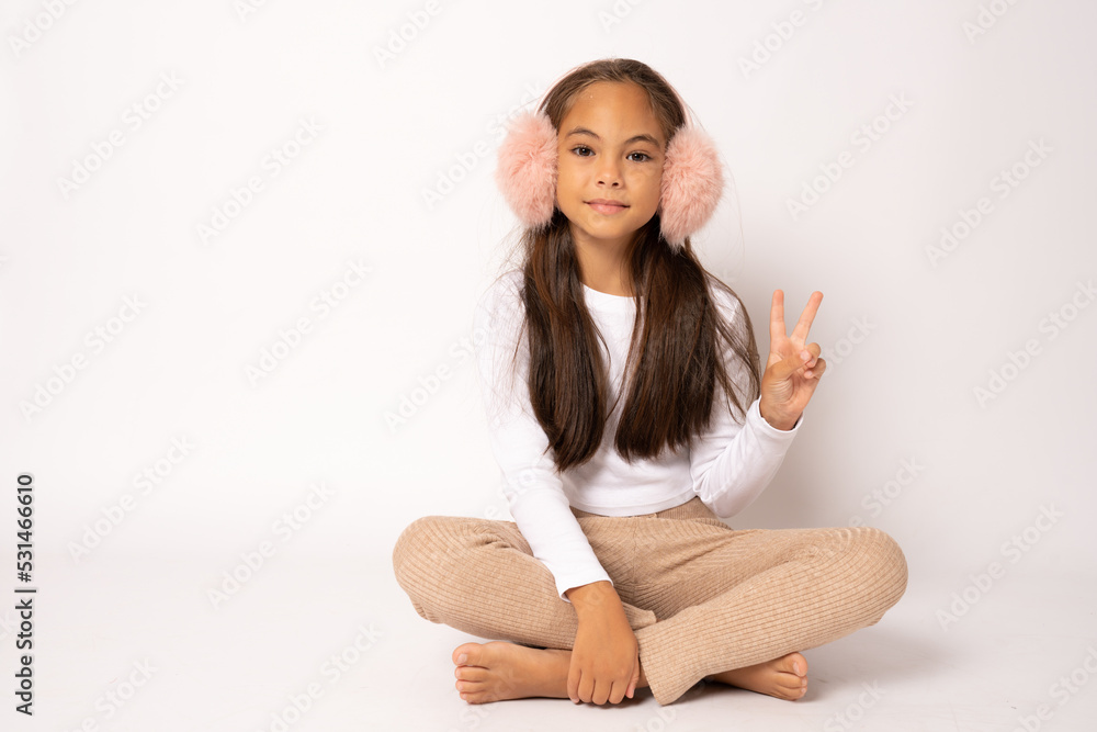 Full length portrait of brunette hispanic girl in earmuffs showing victory sign sitting on floor smiling confident and happy. Full length portrait of brunette hispanic girl in earmuffs showing victory sign sitting on floor smiling confident and happy.