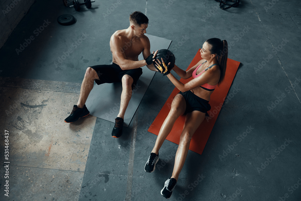 Top view of young fit couple exercising with medicine ball while sitting on exercise mats in gym - obrazy, fototapety, plakaty 