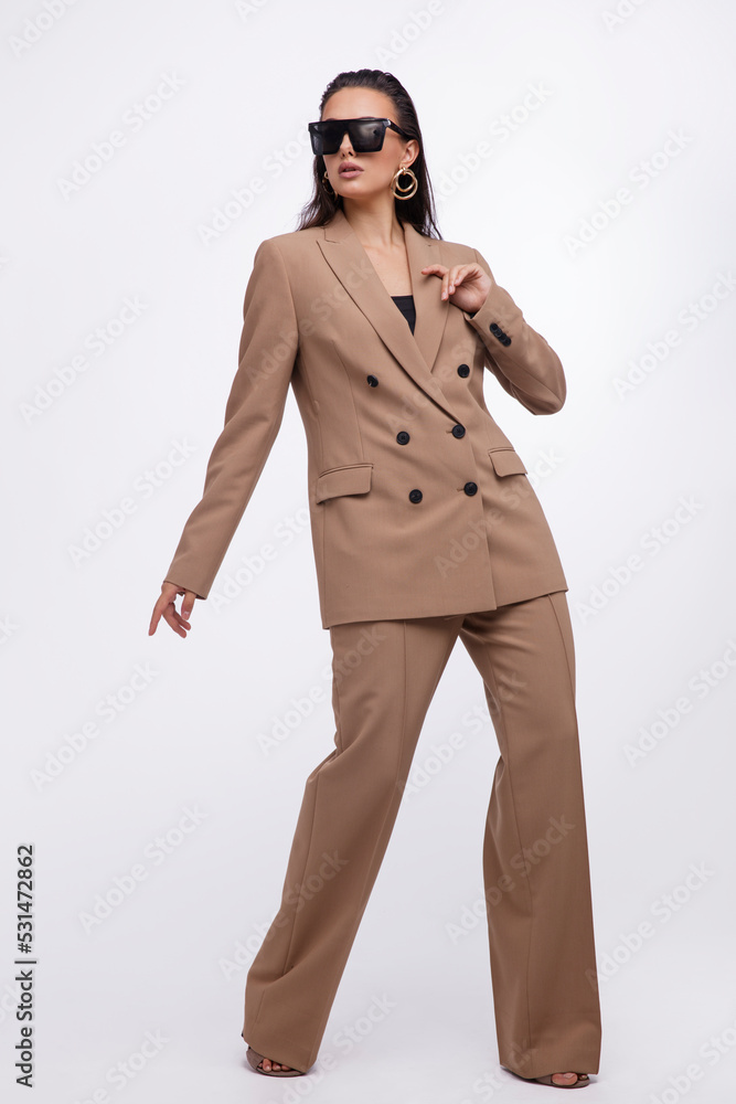 Fashion photo of a beautiful elegant young woman in a pretty oversize brown and beige suit, jacket, pants, stylish sunglasses posing over white background. The hair is gathered back, dark brunette. Fashion photo of a beautiful elegant young woman in a pretty oversize brown and beige suit, jacket, pants, stylish sunglasses posing over white background. The hair is gathered back, dark brunette.