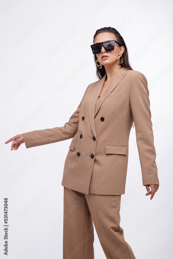 Fashion photo of a beautiful elegant young woman in a pretty oversize brown and beige suit, jacket, pants, stylish sunglasses posing over white background. The hair is gathered back, dark brunette. Fashion photo of a beautiful elegant young woman in a pretty oversize brown and beige suit, jacket, pants, stylish sunglasses posing over white background. The hair is gathered back, dark brunette.