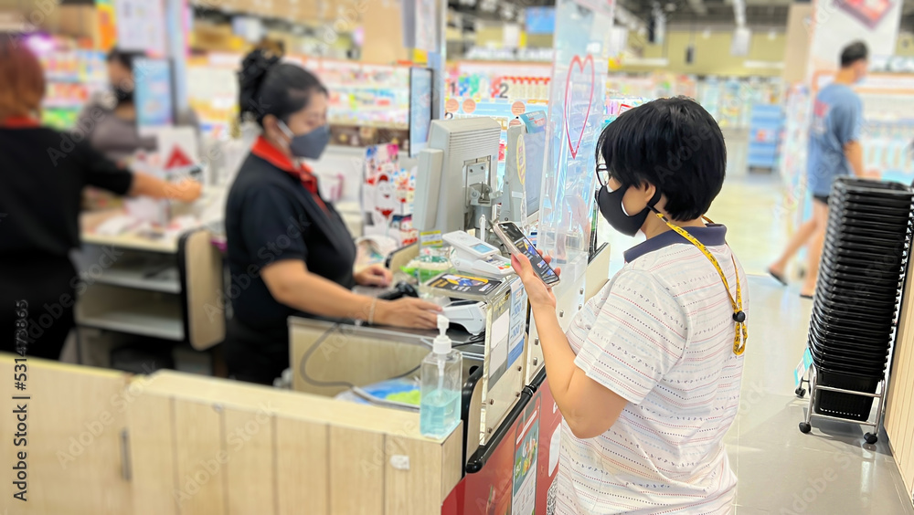 Asian woman wearing a mask while using mobile phone to make contactless payments at supermarket cashier position.