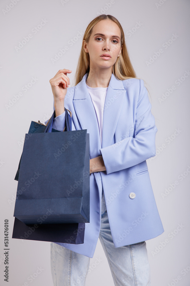 High fashion photo of a beautiful elegant young woman in a pretty blue jacket, blazer, denim jeans, shopping bags posing over white, soft gray background. Studio Shot. Slim Figure, Blonde High fashion photo of a beautiful elegant young woman in a pretty blue jacket, blazer, denim jeans, shopping bags posing over white, soft gray background. Studio Shot. Slim Figure, Blonde