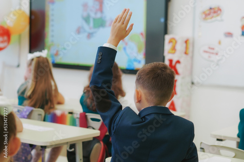 children in school uniforms sitting by desks. Boy raising hand to answer
