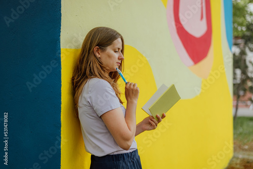 a young woman stands on the street, leaning her back against a multi-colored wall on the street in summer, with a pen and a notebook in her hands, thinking and writing down something