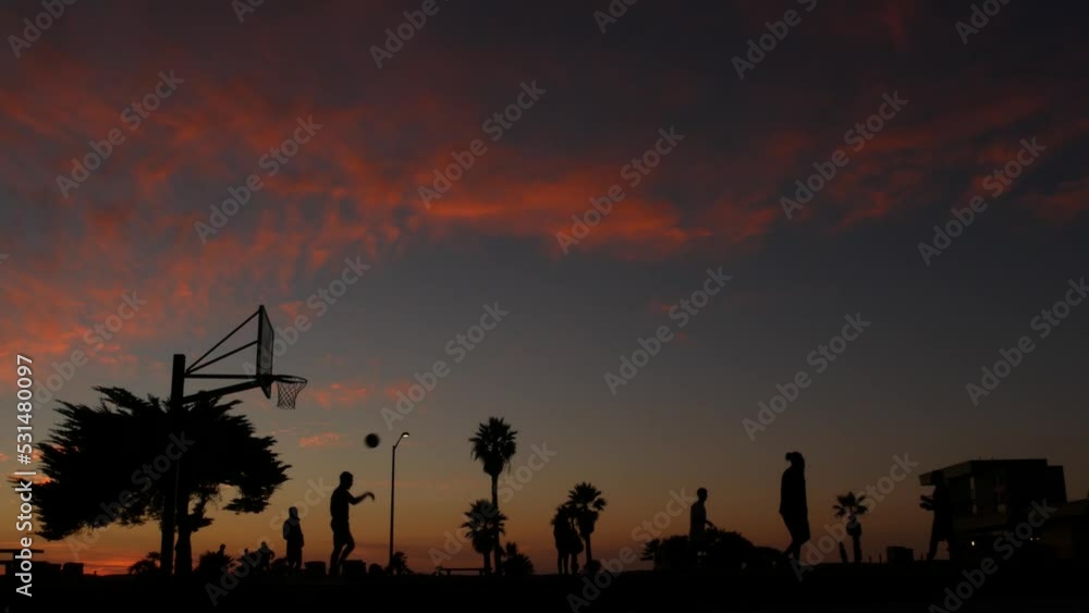 People playing basket ball game, silhouettes of players on basketball court outdoor, sunset ocean beach, California coast, Mission beach, USA. Black hoop, net and backboard on streetball sport field. People playing basket ball game, silhouettes of players on basketball court outdoor, sunset ocean beach, California coast, Mission beach, USA. Black hoop, net and backboard on streetball sport field.
