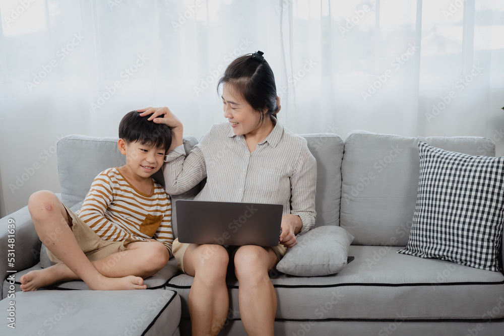 A middle-aged Asian single mom is working on a laptop on the sofa in the living room of the house. The son ran and jumped onto the sofa and sat next to his mother, begging her for attention. A middle-aged Asian single mom is working on a laptop on the sofa in the living room of the house. The son ran and jumped onto the sofa and sat next to his mother, begging her for attention.