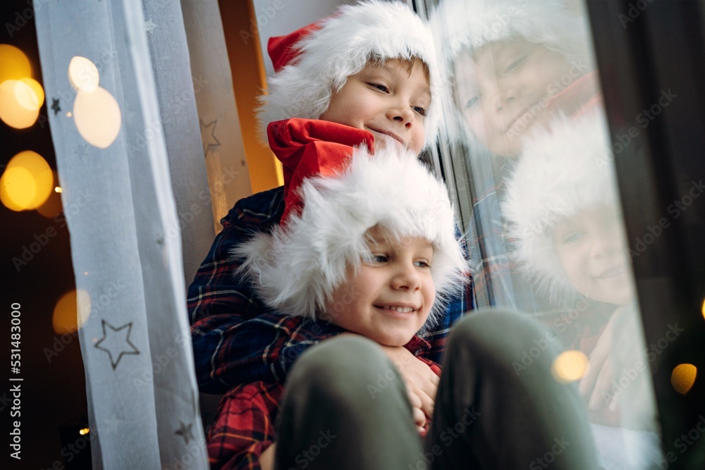 cute children wearing santa hats sitting hugging by the window waitinfg for christmas