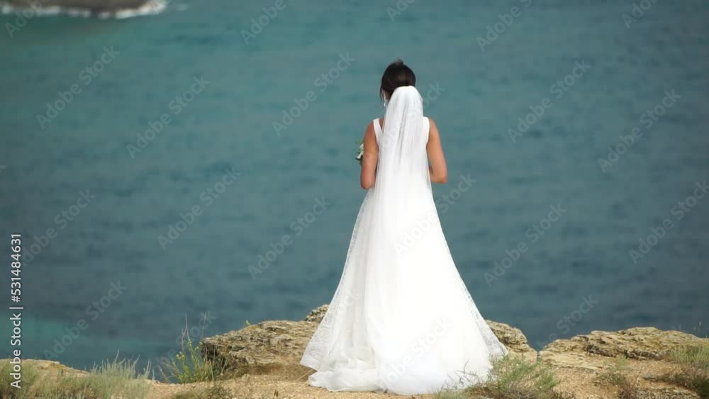 A beautiful bride stands with her back against the background of the sea with a bouquet of flowers. A woman in a long white dress. A beautiful bride stands with her back against the background of the sea with a bouquet of flowers. A woman in a long white dress.