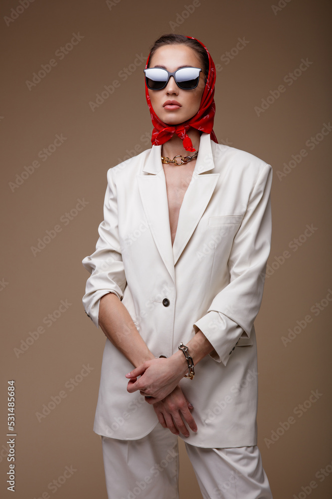 High fashion photo of a beautiful elegant young woman in pretty white suit, jacket, trousers, pants, shoes, red scarf, sunglasses, accessories, posing on beige  background. Studio Shot. Portrait.