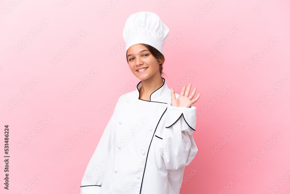 Little caucasian chef girl isolated on pink background saluting with hand with happy expression