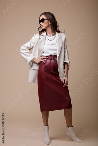 High fashion photo of a beautiful elegant young woman in a pretty white leather jacket, red long skirt, boots, accessories, stylish sunglasses posing over beige background. Studio Shot. Portrait.