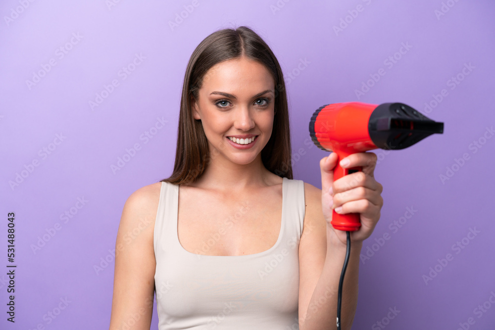 Young caucasian woman holding a hairdryer isolated on purple background with happy expression Young caucasian woman holding a hairdryer isolated on purple background with happy expression