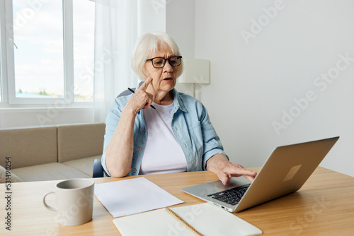 an attentive, focused elderly businesswoman is sitting at home in a bright interior at work at a laptop and, while conducting video training, looks attentively, holding her index finger near her face