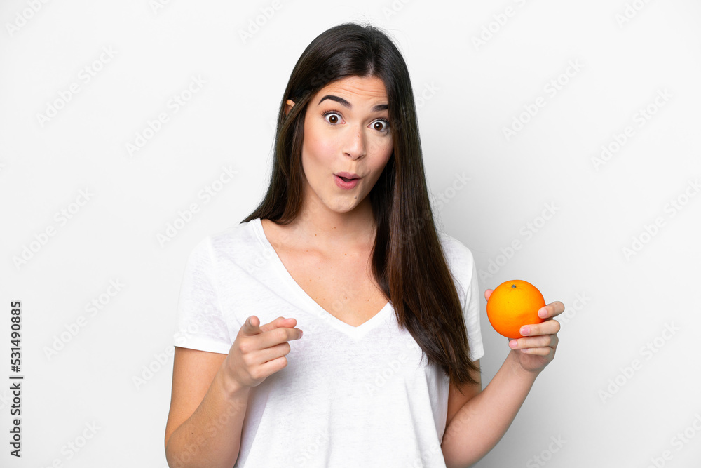 Young Brazilian woman holding an orange isolated on white background surprised and pointing front Young Brazilian woman holding an orange isolated on white background surprised and pointing front