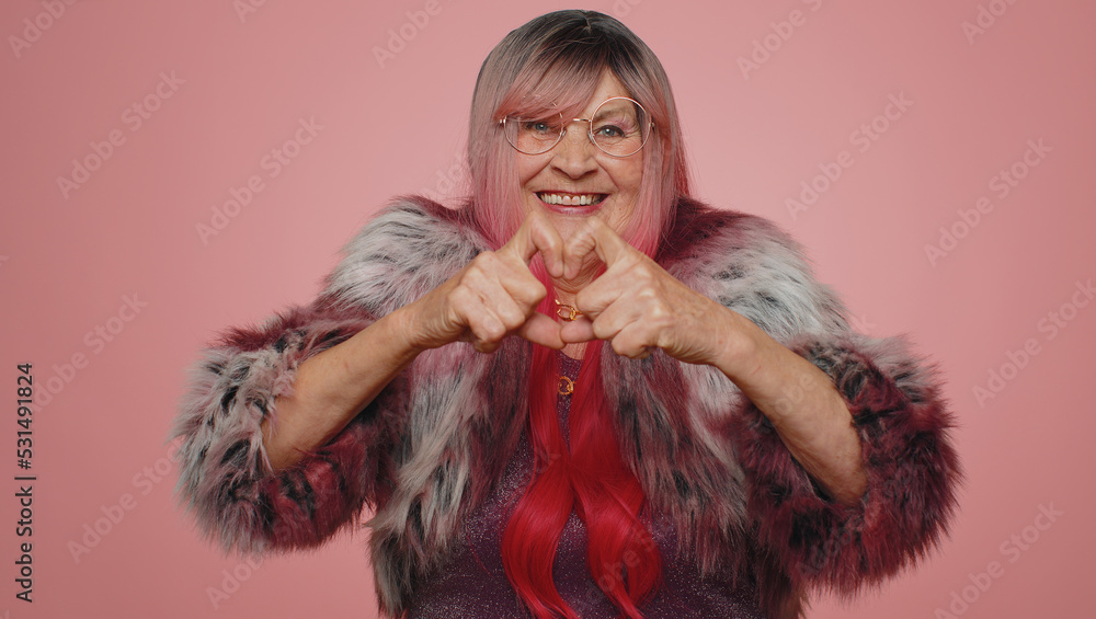Woman in love. Smiling stylish old woman makes heart gesture demonstrates love sign expresses good feelings and sympathy. Senior elderly grandmother 70s isolated alone on pink studio wall background