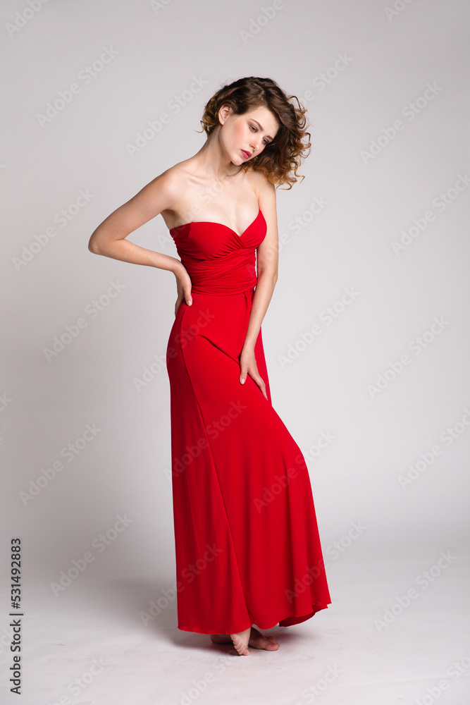 High fashion photo of a beautiful elegant young woman in a pretty long red dress, hairstyle on white, soft gray background. Studio Shot. 