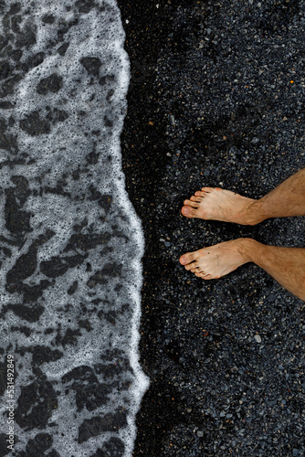 Legs on the background of the beach. Top view of bare feet and legs in fine black pebbles with the movement of waves coming to the feet - foaming sea texture. The concept of summer holidays and vacati