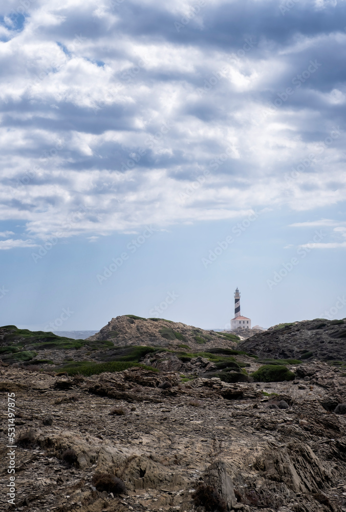 distant image of the Favaritx lighthouse, with a rock formation around it on a cloudy summer day, vertical, copy space, Menorca, Balearic Islands, spain