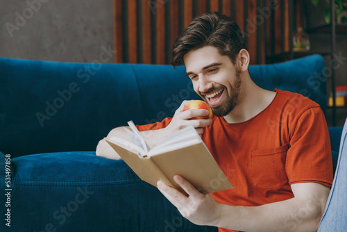 Young calm man 20s wears red t-shirt bite apple read book novel sit on blue sofa couch stay at home hotel flat rest relax spend free spare time in living room indoors grey wall. People lounge concept. photo