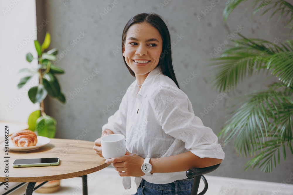 Side view young fun calm tranquil latin woman 30s wear white shirt hold cup drink coffee look aside sit alone at table in coffee shop cafe restaurant indoors. Freelance mobile office business concept. Side view young fun calm tranquil latin woman 30s wear white shirt hold cup drink coffee look aside sit alone at table in coffee shop cafe restaurant indoors. Freelance mobile office business concept.