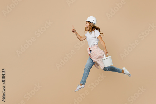 Full size young employee laborer handyman woman in white t-shirt helmet jump high hold paint roller bucket point aside isolated on plain beige background Instruments renovation room Repair concept.