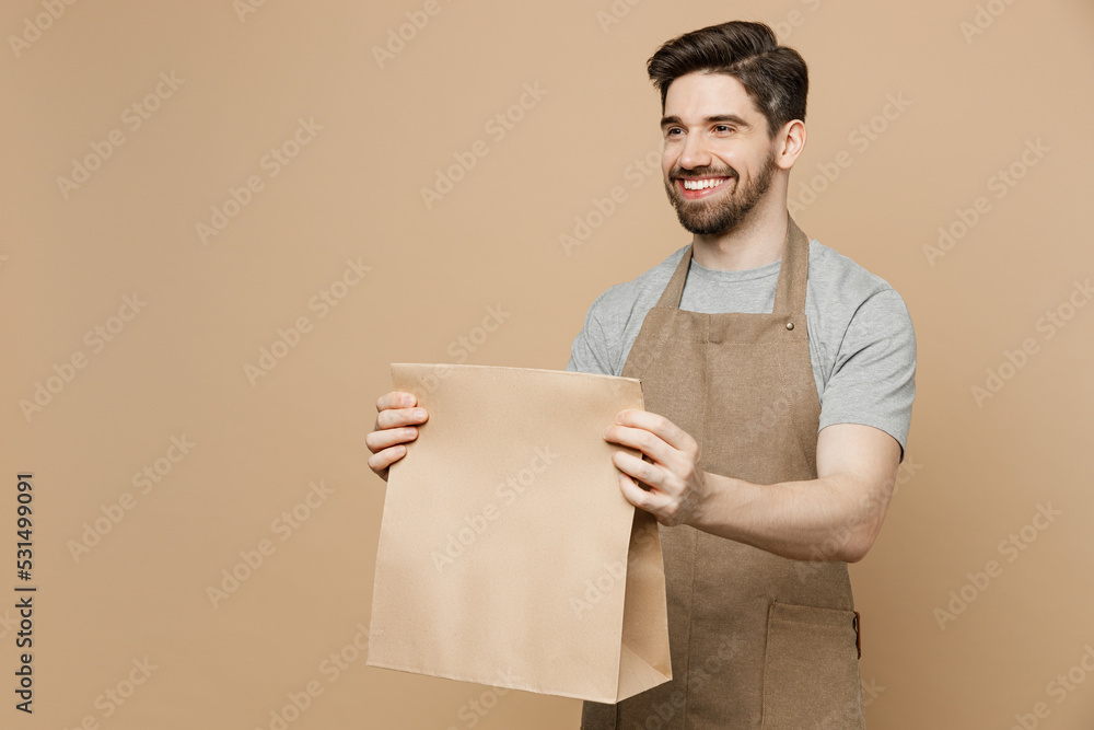 Young man barista barman employee wear brown apron work in coffee shop give craft paper bag for takeaway food products isolated on plain pastel light beige background. Small business startup concept. Young man barista barman employee wear brown apron work in coffee shop give craft paper bag for takeaway food products isolated on plain pastel light beige background. Small business startup concept.