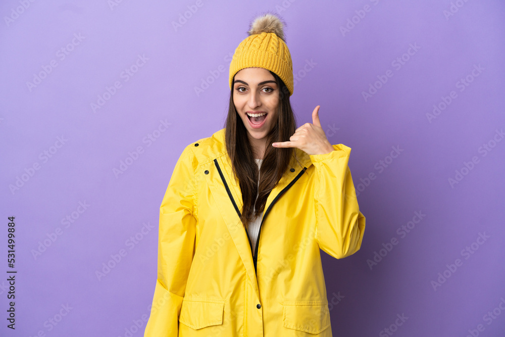 Young caucasian woman wearing a rainproof coat isolated on purple background making phone gesture. Call me back sign Young caucasian woman wearing a rainproof coat isolated on purple background making phone gesture. Call me back sign