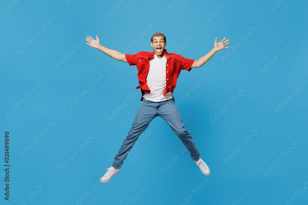 Full size young fun excited man of African American ethnicity 20s wear red shirt jump high with outstretched hands legs isolated on plain pastel light blue cyan background. People lifestyle concept