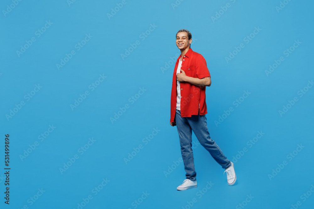 Full body side view happy young man of African American ethnicity 20s he wear red shirt look camera strolling walk going isolated on plain pastel light blue cyan background. People lifestyle concept.