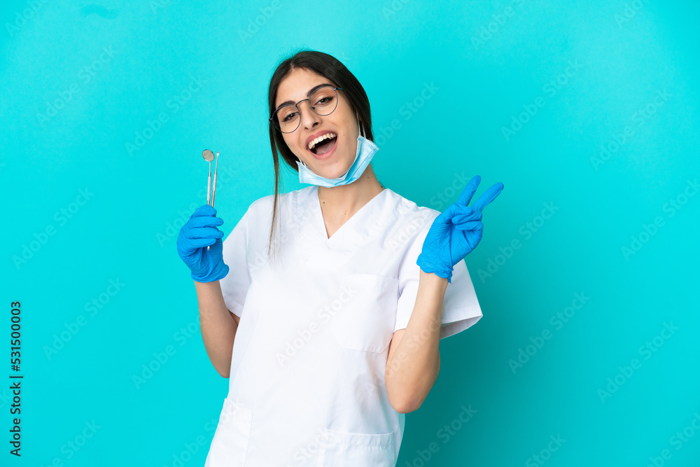 Young caucasian dentist woman holding tools isolated on blue background smiling and showing victory sign Young caucasian dentist woman holding tools isolated on blue background smiling and showing victory sign