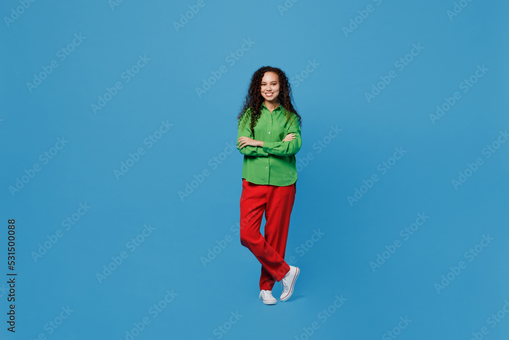 Full body young smiling woman of African American ethnicity 20s she wear green shirt hold hands crossed folded look camera isolated on plain blue background studio portrait. People lifestyle concept.