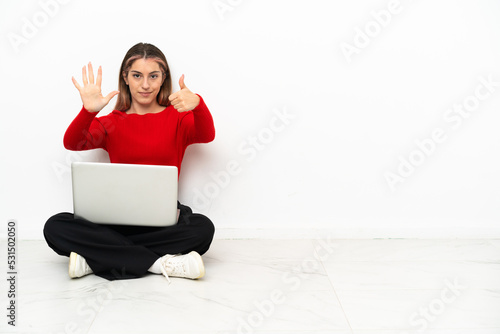 Young caucasian woman with a laptop sitting on the floor counting six with fingers