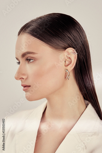 Cropped close-up shot of a young woman with two asymmetrical golden snake ear cuffs. A dark-haired woman with crystal snake ear cuffs is on a pastel background. Side view.