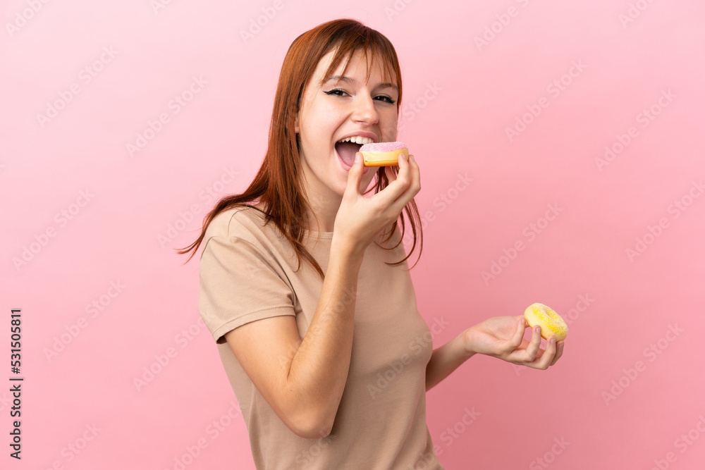 Redhead girl isolated on pink background eating a donut Redhead girl isolated on pink background eating a donut