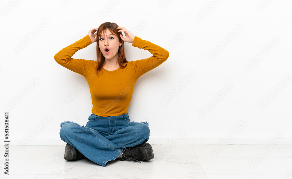 Redhead girl sitting on the floor isolated on white background with surprise expression Redhead girl sitting on the floor isolated on white background with surprise expression