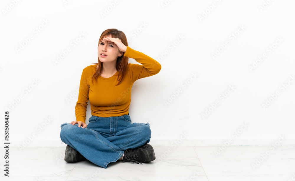 Redhead girl sitting on the floor isolated on white background looking far away with hand to look something Redhead girl sitting on the floor isolated on white background looking far away with hand to look something