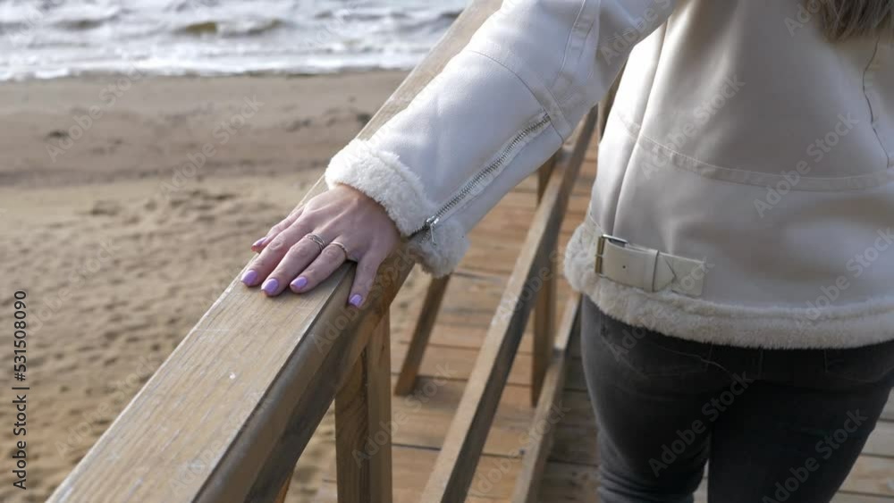 A girl in jeans and a jacket walks along a wooden pier, putting her hand on the railing, a view from the back. A girl in jeans and a jacket walks along a wooden pier, putting her hand on the railing, a view from the back.