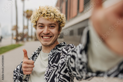 Smiling young caucasian guy takes picture of himself and shows class sign. Curly hair man dressed in casual clothes outdoor. Concept self shooting.