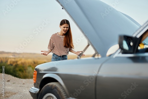 A sad woman has opened the hood of a broken down car and is looking for the cause of the breakdown on a road trip alone with her hands spread apart and shouting in anger © SHOTPRIME STUDIO