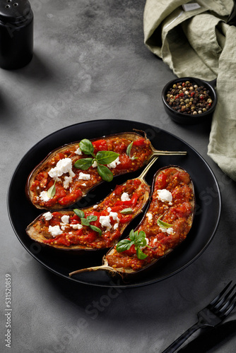 Eggplants stuffed with red sauce, chopped tomatoes, sweet pepper, feta cheese and basil on black plate and dark background.