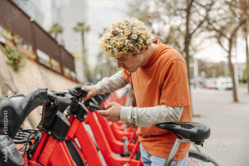 Curly young caucasian guy rents bicycle through app on his phone. Blonde male is active lifestyle. Concept sport, gadgets, apps.