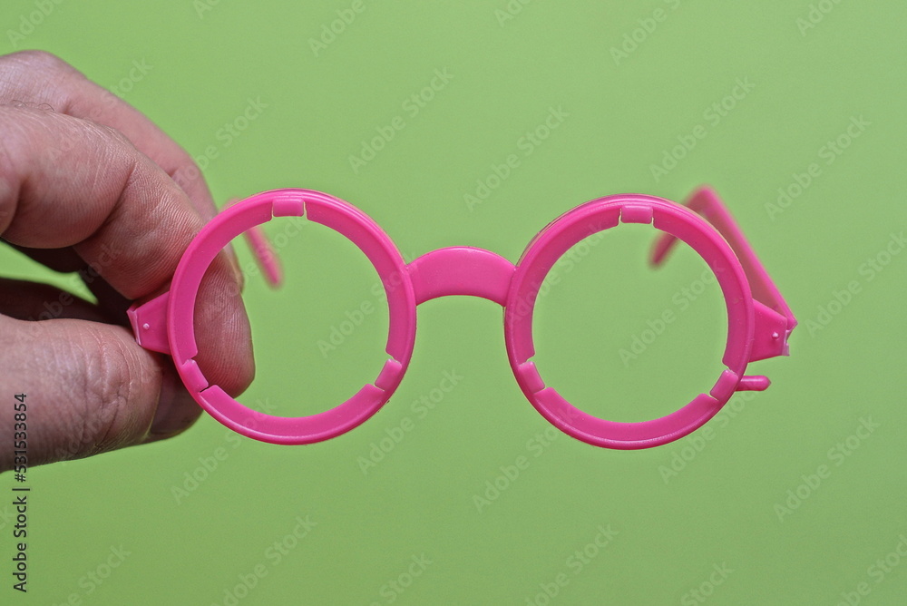 Fototapeta premium fingers on the hand hold one plastic pink toy glasses on a green background