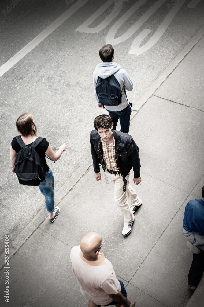 Young European man, wearing black jacket, light color pants, walking on sidewalk on street in New York City, passing by people, looking around.